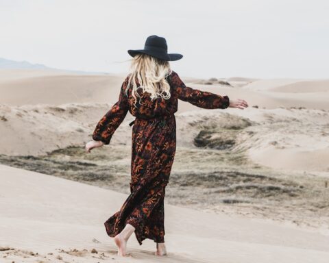 photo of person wearing brown and orange floral maxi dress walking barefooted along deserted land